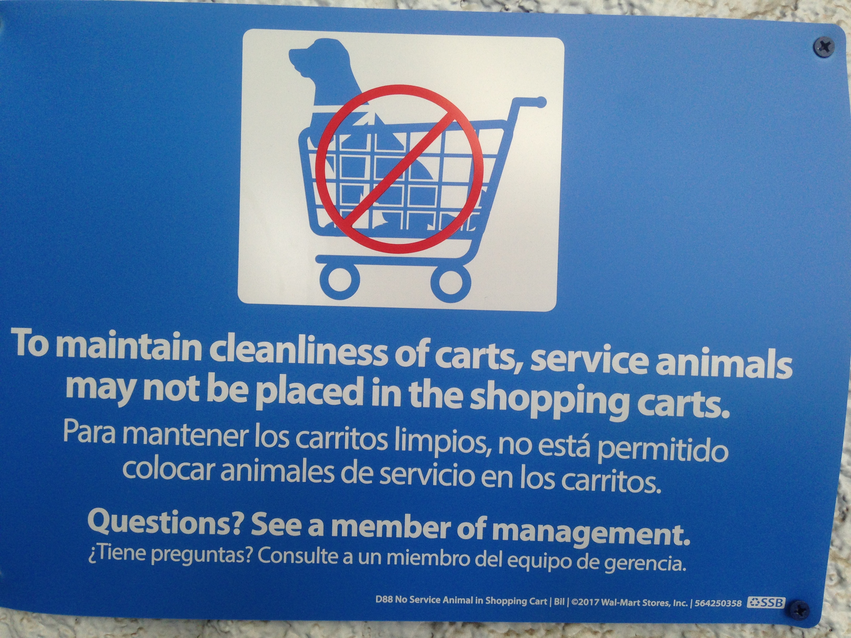 A line illustration of a dog sitting in a shopping cart facing forward, with the red “no” symbol over it. Beneath the subtext in English and Spanish reads: To maintain cleanliness of carts, service animals may not be placed in the shopping carts. / Questions? See a member of management.