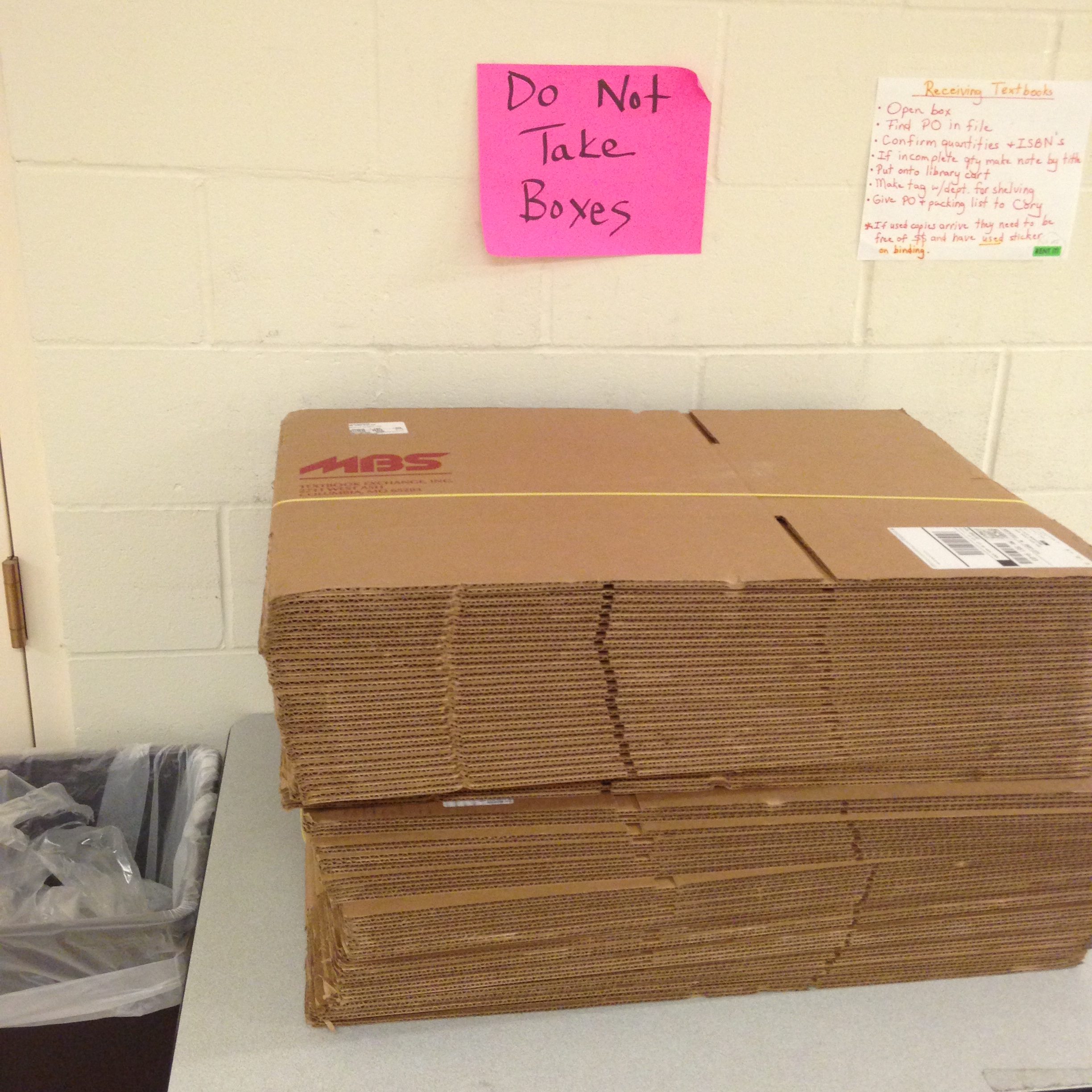 A handwritten sign in chisel-point Sharpie on pink construction paper, reading “Do Not Take Boxes”, affixed to a cinder-block wall above a table bearing several sets of ten new, flattened cardboard boxes still zip-tied together.