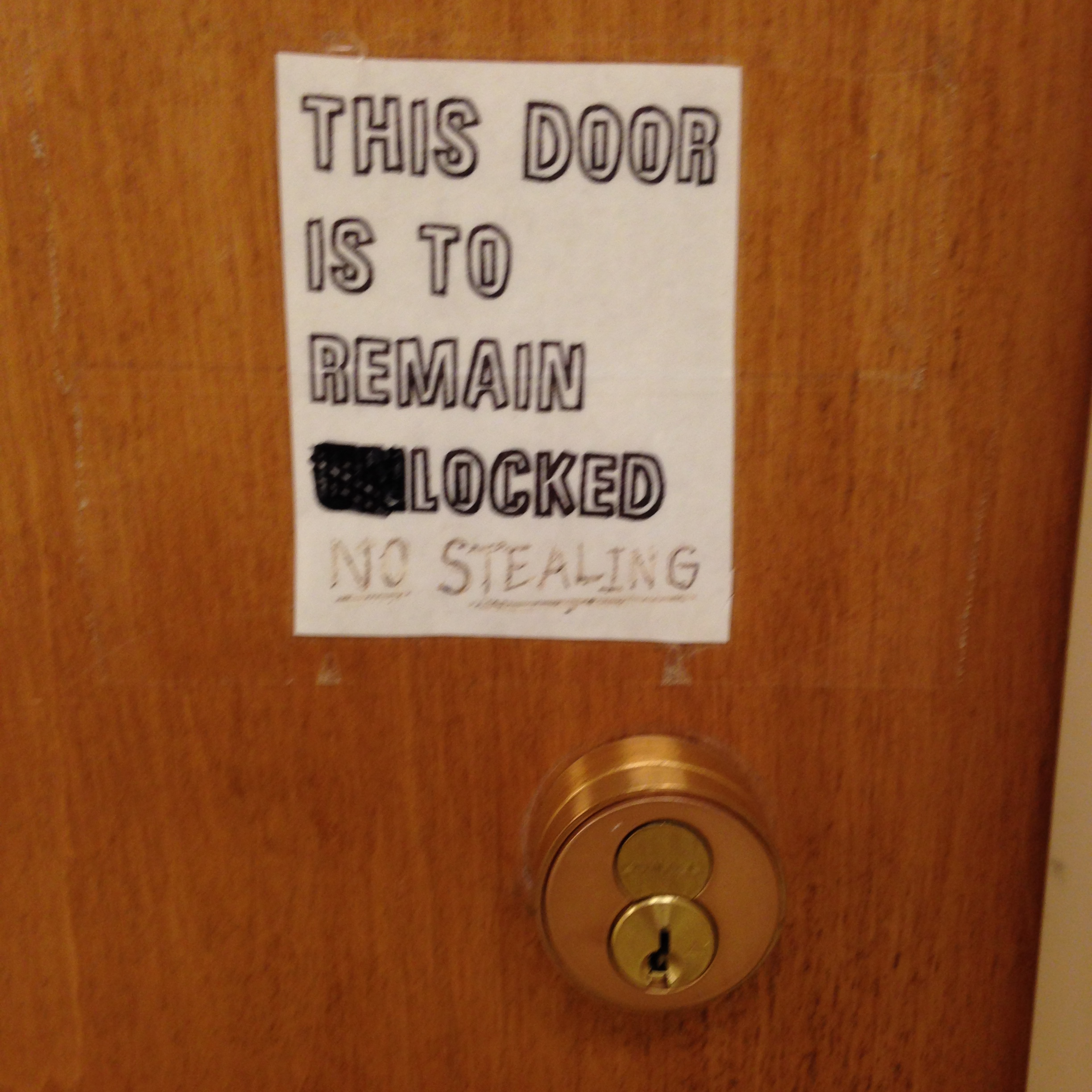 A deadbolt cylinder on the right side of a door, with a sign above it reading in all-caps stenciled letters, “This Door Is To Remain Unlocked.” The “UN” in “Unlocked” has been blacked out with a marker, and underneath someone has written in underlined text in a marker that didn’t apply to the surface well, “No Stealing.”