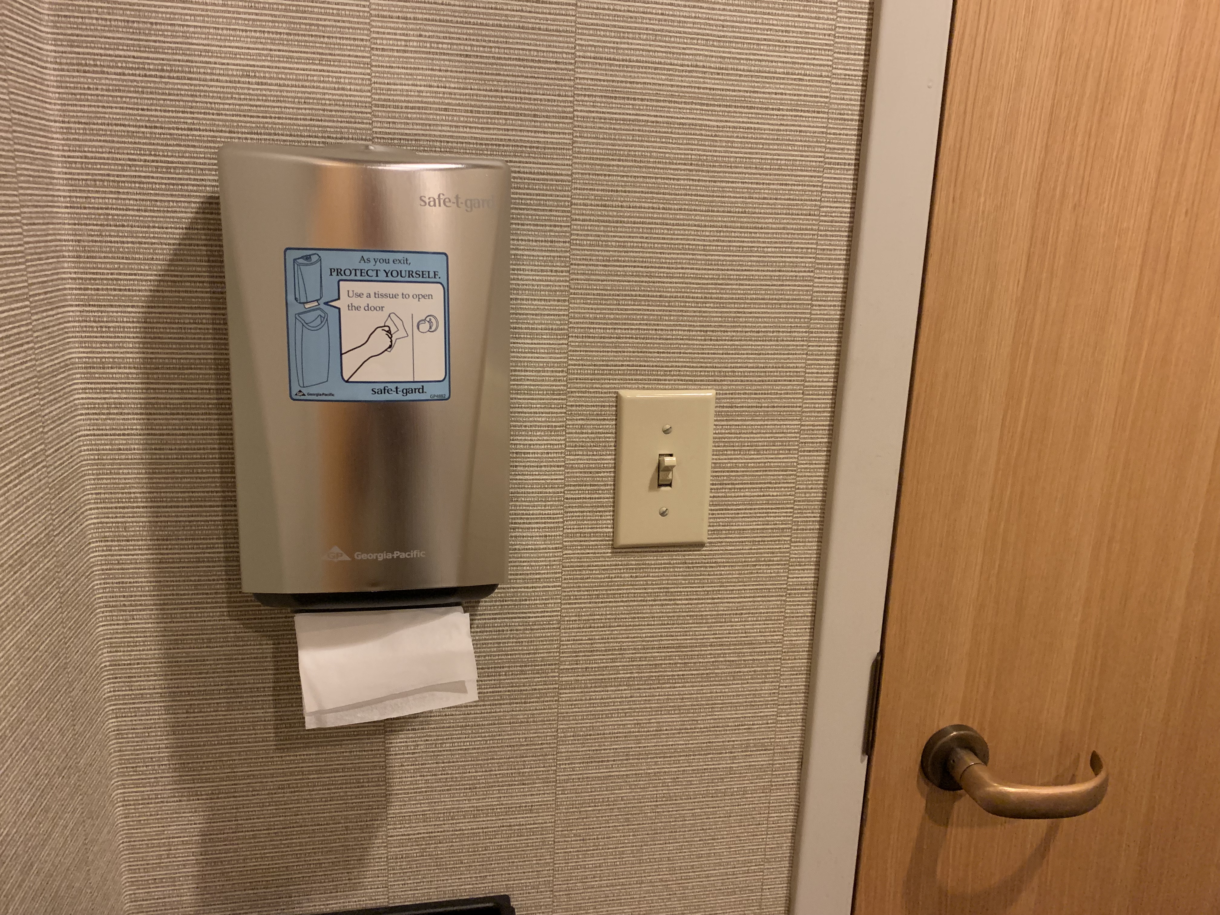 On the right side of the picture, a door with door handle opening inwards. On the left side, a light switch, and to its left a small paper-towel dispenser of sorts dispensing single tissues from the bottom, with the outline of a trash can in the picture below. A sign on the dispenser reads: “As you exit, PROTECT YOURSELF. Use a tissue to open the door.” The second sentence is shown inside a speech bubble emanating from a picture of the dispenser. Fortunately, the picture of the dispenser on the dispenser does not contain a picture of the dispenser to prevent infinite regress.